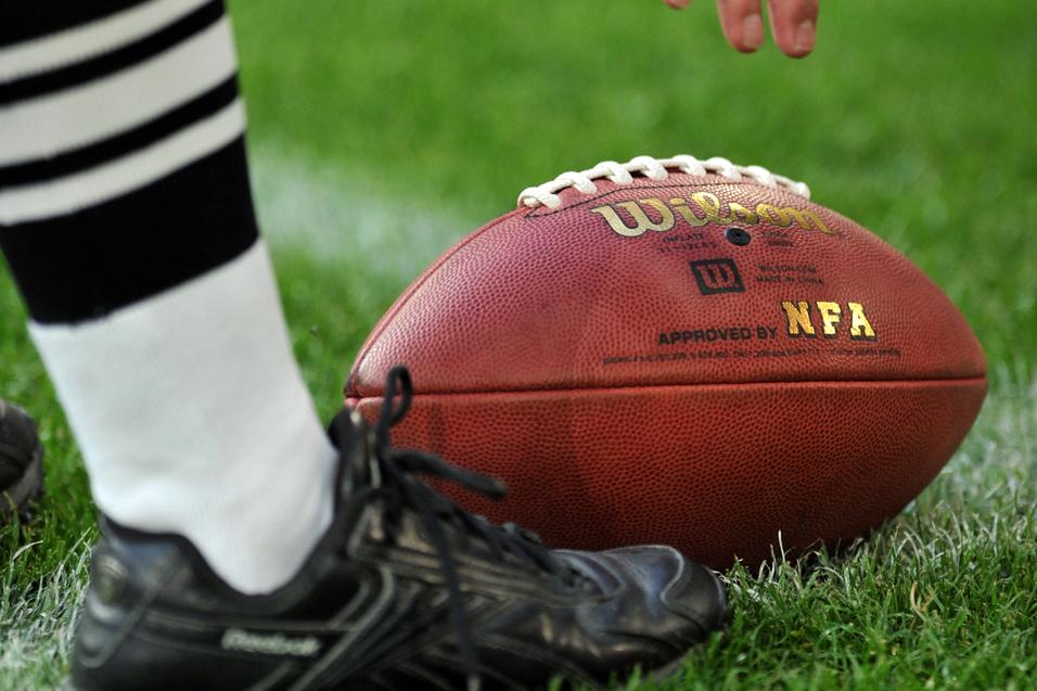 Nahaufnahme eines American Footballs auf dem Gras, daneben ein Schuh und eine Hand, die den Ball berührt.