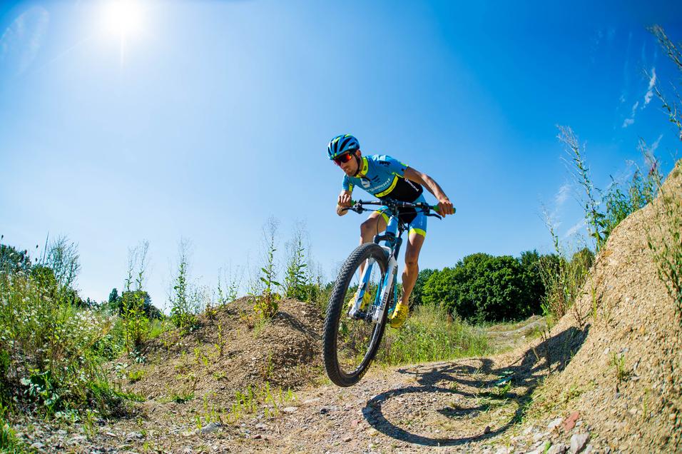 Mountainbiker springt über einen Erdhügel auf einem sonnigen, grasbewachsenen Trail.