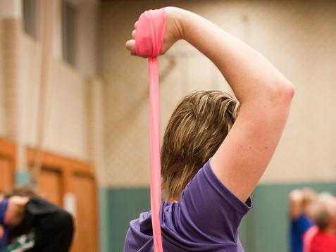 Frau in lila T-Shirt dehnt mit einem pinken Widerstandsband ihren Arm in einer Sporthalle. Hintergrund mit anderen Personen.