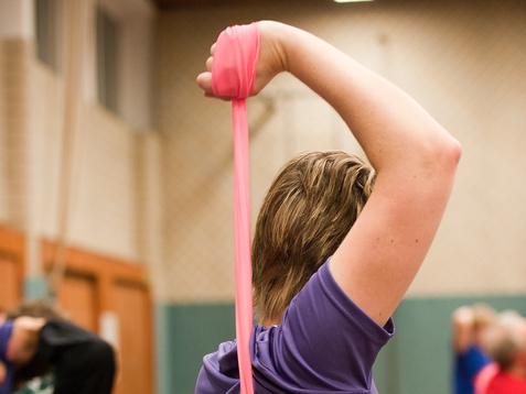 Frau in lila T-Shirt dehnt mit einem pinken Widerstandsband ihren Arm in einer Sporthalle. Hintergrund mit anderen Personen.