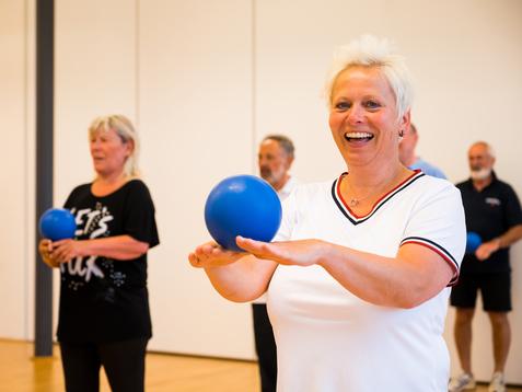 Gruppe älterer Menschen bei einer Übung mit blauen Bällen in einem hellen Raum. Fokus auf lächelnde Frau vorne.