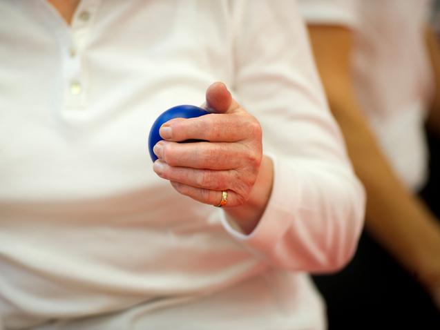 Nahaufnahme einer Hand, die einen blauen Ball hält, während die Person in einem weißen Oberteil sitzt.