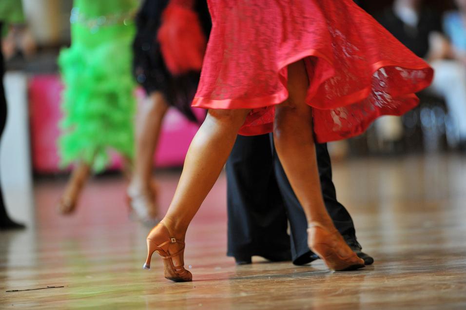 Tanzende Beine in eleganten Schuhen, während sich ein rotes Kleid dreht, vor unscharfem Hintergrund.