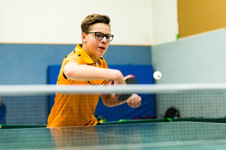 Junger Tischtennisspieler in orangefarbenem Shirt schlägt den Ball auf einem Tischtennistisch.
