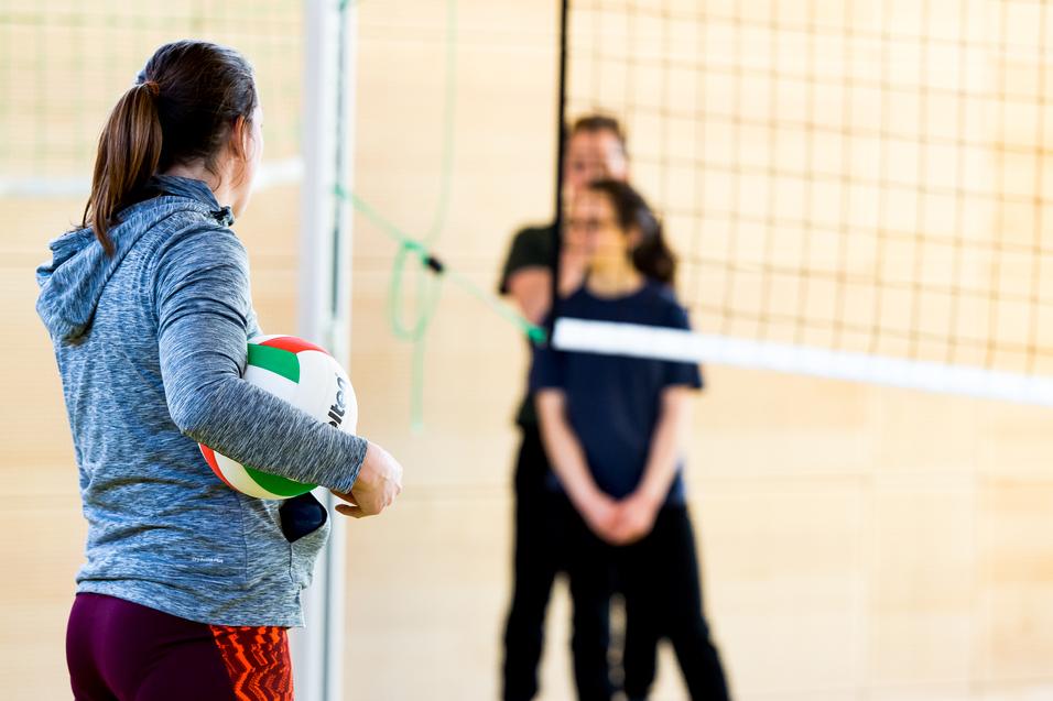 Eine Person mit einem Volleyball steht vor dem Netz, während zwei weitere Personen im Hintergrund warten.